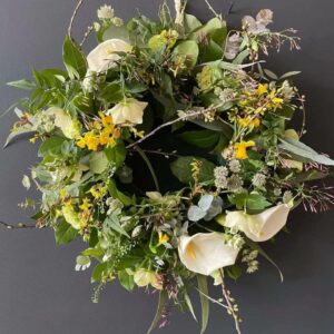 The photo showcases an floral wreath, with beautiful foliage and florals. Wreath is sat on a dark grey wall to show contrast.